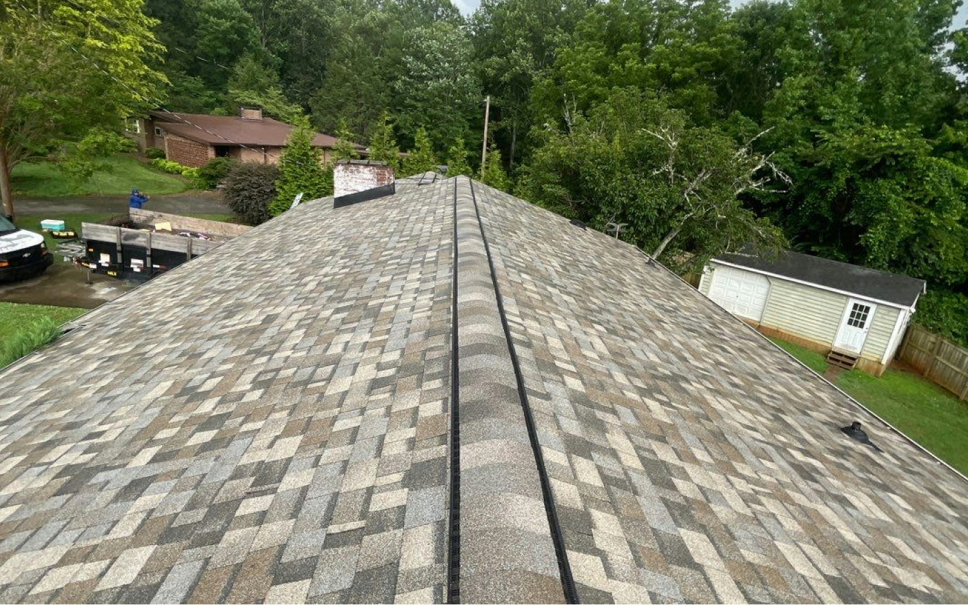 A new gray, tan, and brown shingle roof on a house, with green trees and a cloudy sky in the background.