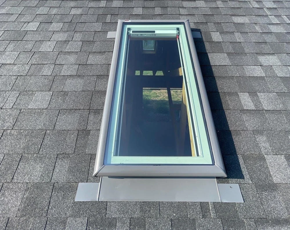 A newly installed rectangular skylight on a dark gray shingled roof under a clear blue sky
