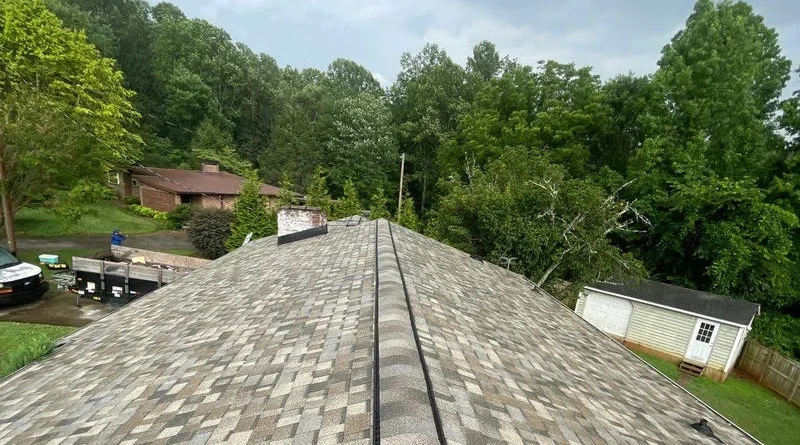 A new gray, tan, and brown shingle roof on a house, with green trees and a cloudy sky in the background.